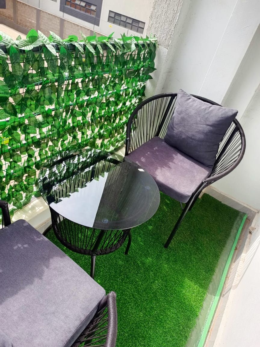 Un pequeño espacio al aire libre con una mesa redonda y dos sillas, rodeado de follaje verde artificial.