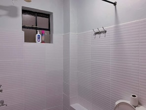 A bathroom with white tiled walls, a window with a shelf holding toiletries, a towel rack, and a toilet. - Amaziah Apartments Muthiga (Rungiri)