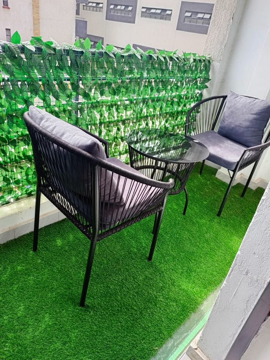 Un balcón con césped artificial verde, dos sillas de metal negro y una mesa de vidrio.