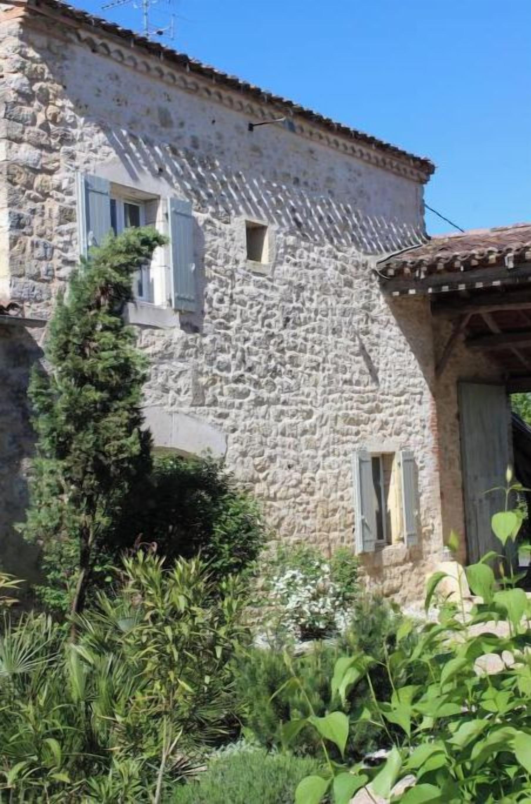 A stone building with a rustic appearance, featuring a small window with shutters and a door with a tiled roof.
