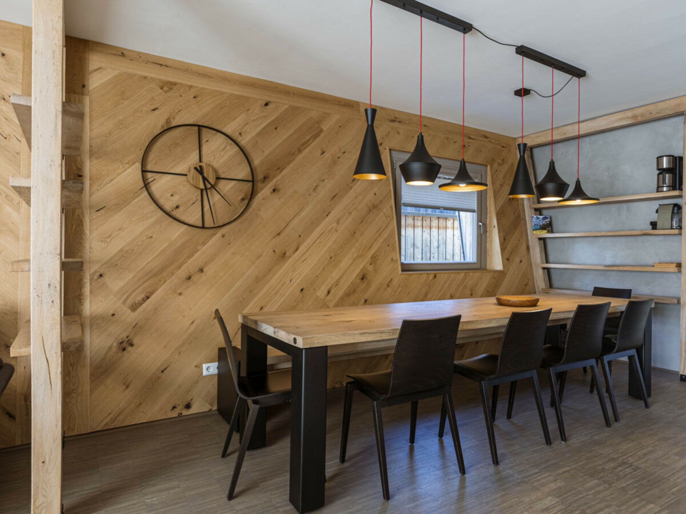 A modern dining area with a wooden herringbone wall, a round wall clock, and pendant lighting.
