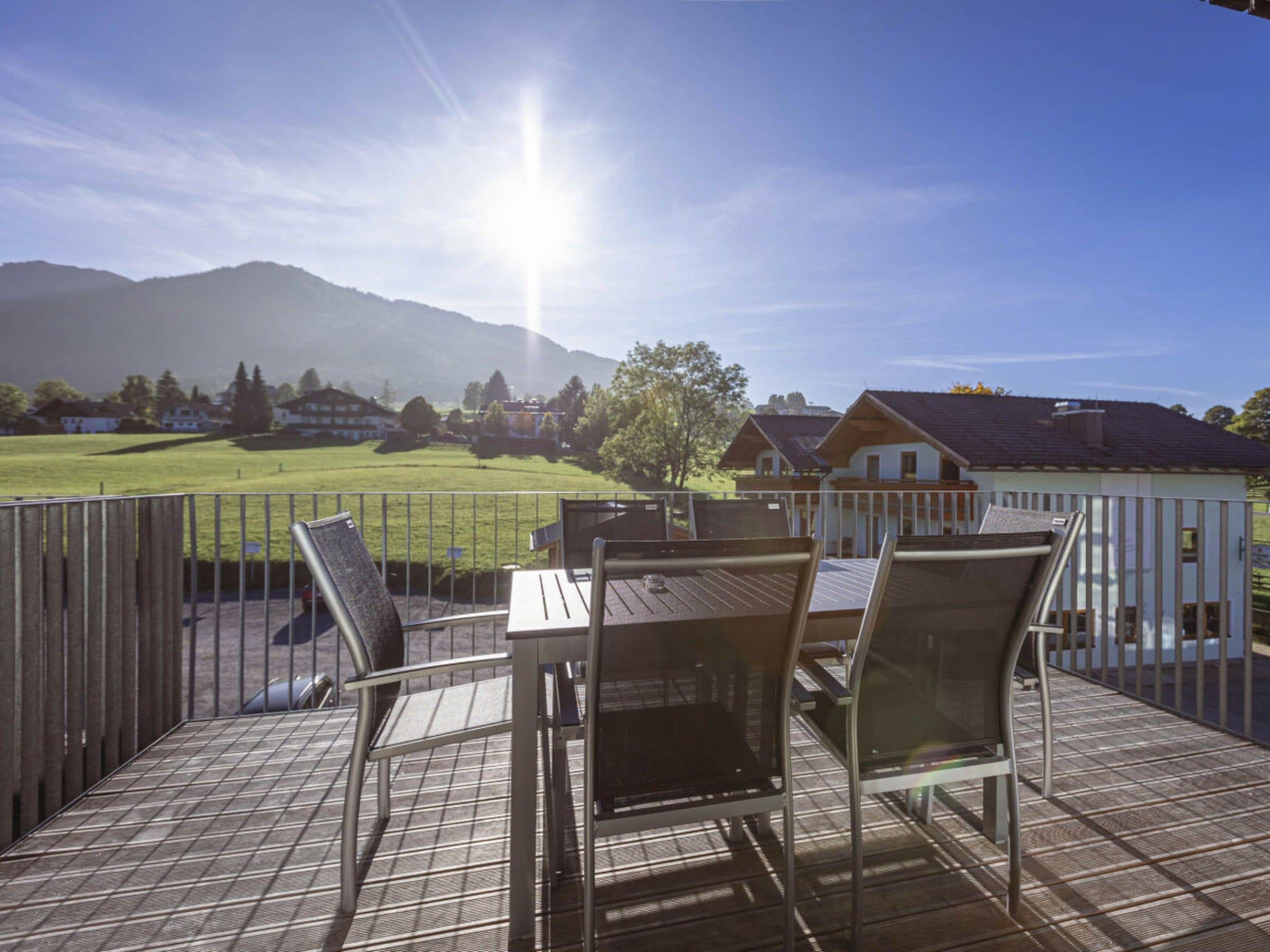An outdoor deck with a table and chairs, overlooking a grassy field and houses with mountains in the background.