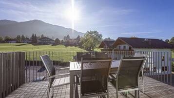 An outdoor deck with a table and chairs, overlooking a grassy field and houses with mountains in the background.