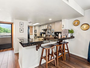 A modern kitchen with a bar area, wooden stools, and decorative wall art. - Kailua-Kona, Holualoa Bay Villas, Spacious 4 BR, Beachfront, Sleeps 12 (Kailua-Kona)
