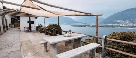 Un banc et une table en pierre, placés sous une pergola en bois, offrent une vue sur un lac, avec des montagnes en arrière-plan.