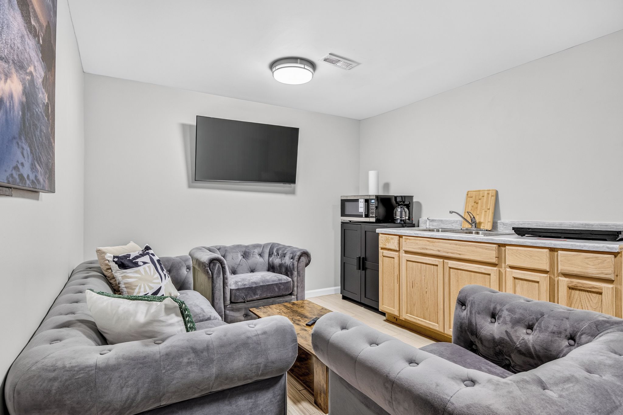 A modern living room with a grey sofa set, a wooden coffee table, and a kitchen area with a microwave and coffee machine.