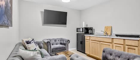 Een moderne woonkamer met een grijze zithoek, een houten salontafel, en een keuken met een magnetron en koffieapparaat.