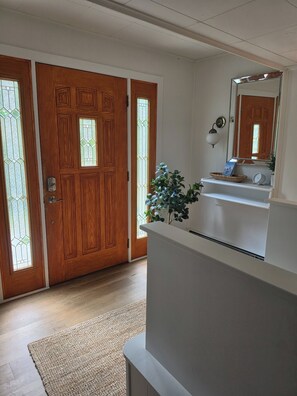 Un pasillo con una puerta de madera, pared blanca, un espejo, una planta y un estante con una cesta y un reloj.