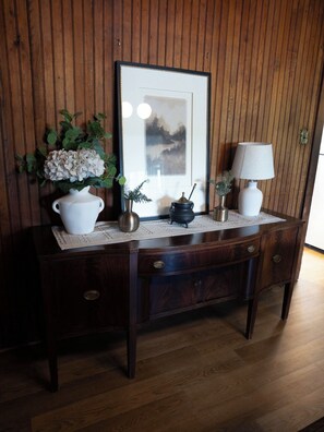 Une commode en bois avec un miroir, un vase blanc avec des fleurs, une petite plante en pot et une lampe.