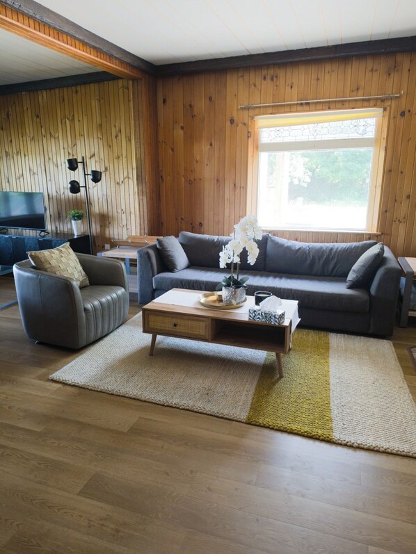 A living room with a sofa set, a coffee table, a television, and a window with a view of greenery.