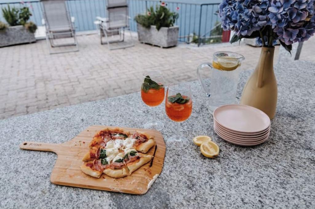 Une pizza sur une planche en bois, deux verres contenant une boisson rouge, un vase avec des fleurs et une pile d’assiettes sur une surface en pierre.