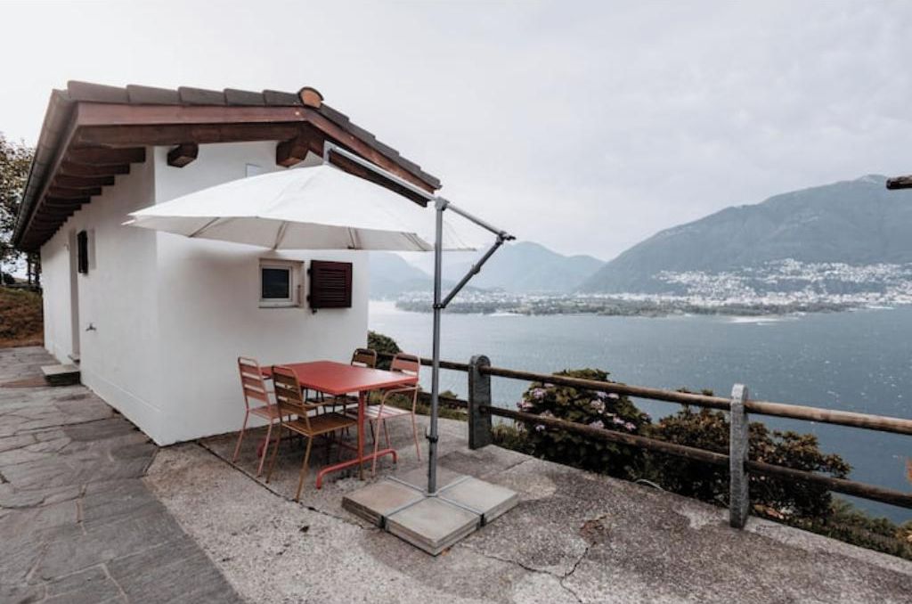 Un pequeño edificio blanco con una mesa y sillas rojas, un paraguas blanco y vistas a un lago con montañas al fondo.