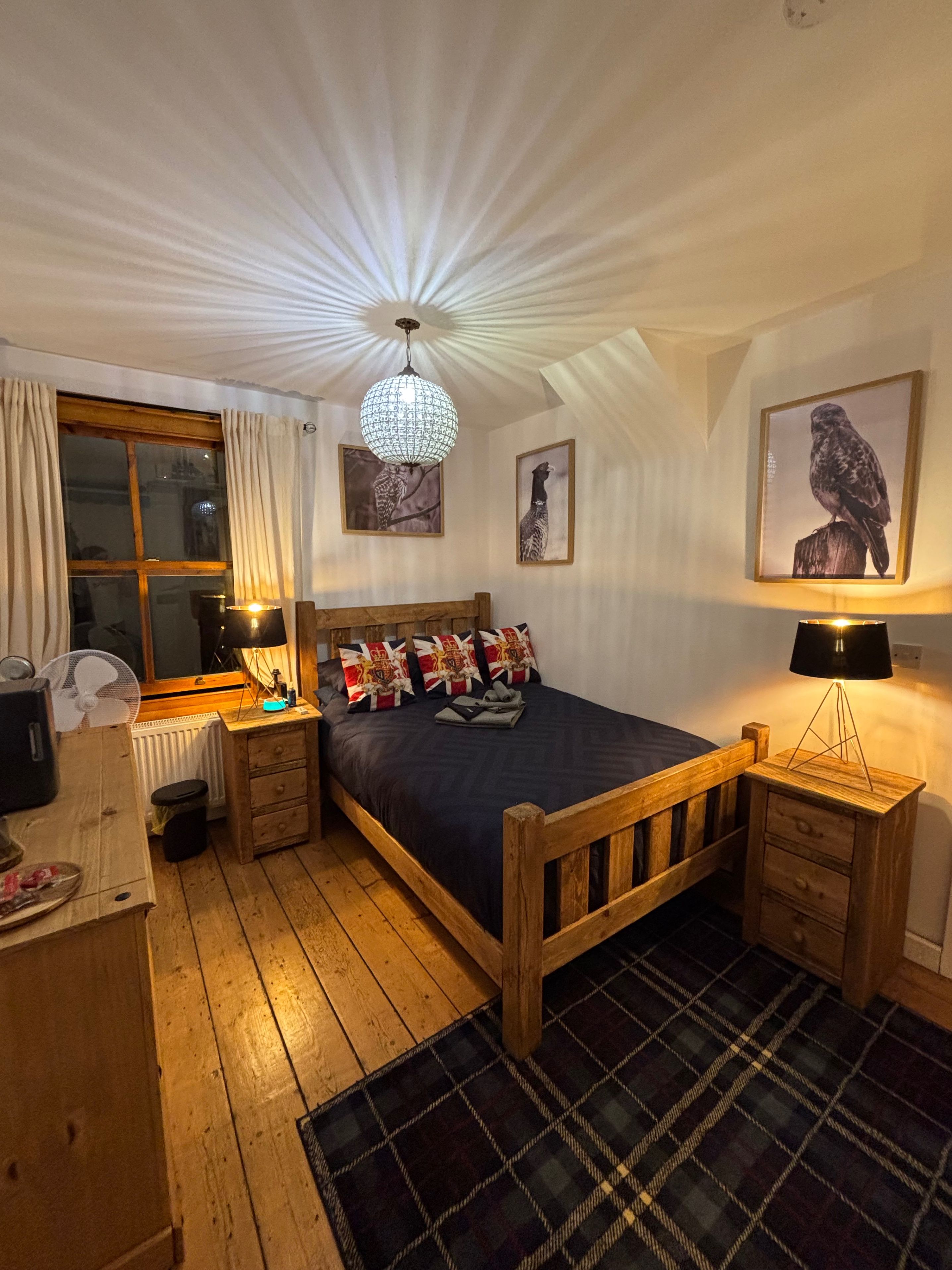 A bedroom with a wooden bed, a nightstand, a lamp, a radiator, a window with curtains, and wall art.