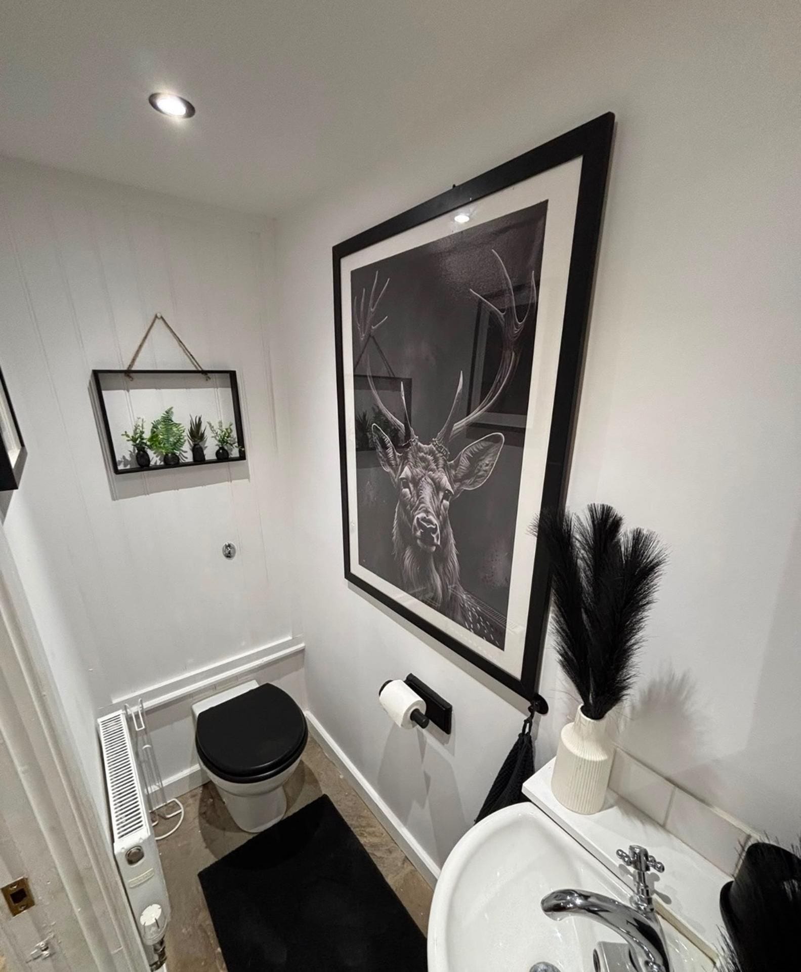 A modern bathroom with a framed deer artwork, a small plant shelf, a toilet, and a sink with a decorative vase.