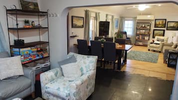 Ein Wohnzimmer mit einer Couch, einem gemusterten Sessel, einem Esstisch mit Stühlen und einem Bücherregal.