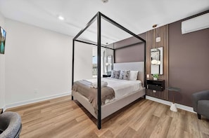 A modern bedroom with a four-poster bed, a bedside table with a lamp, and a view of the outdoors. - The Columbus Hotel - The Reimagined Suite - 52-304 (Middletown)