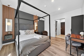 A modern bedroom with a four-poster bed, a wooden accent wall, a bedside table with a lamp, and a view into the adjacent dining area. - The Columbus Hotel - The Market Suite - 52-202 (Middletown)