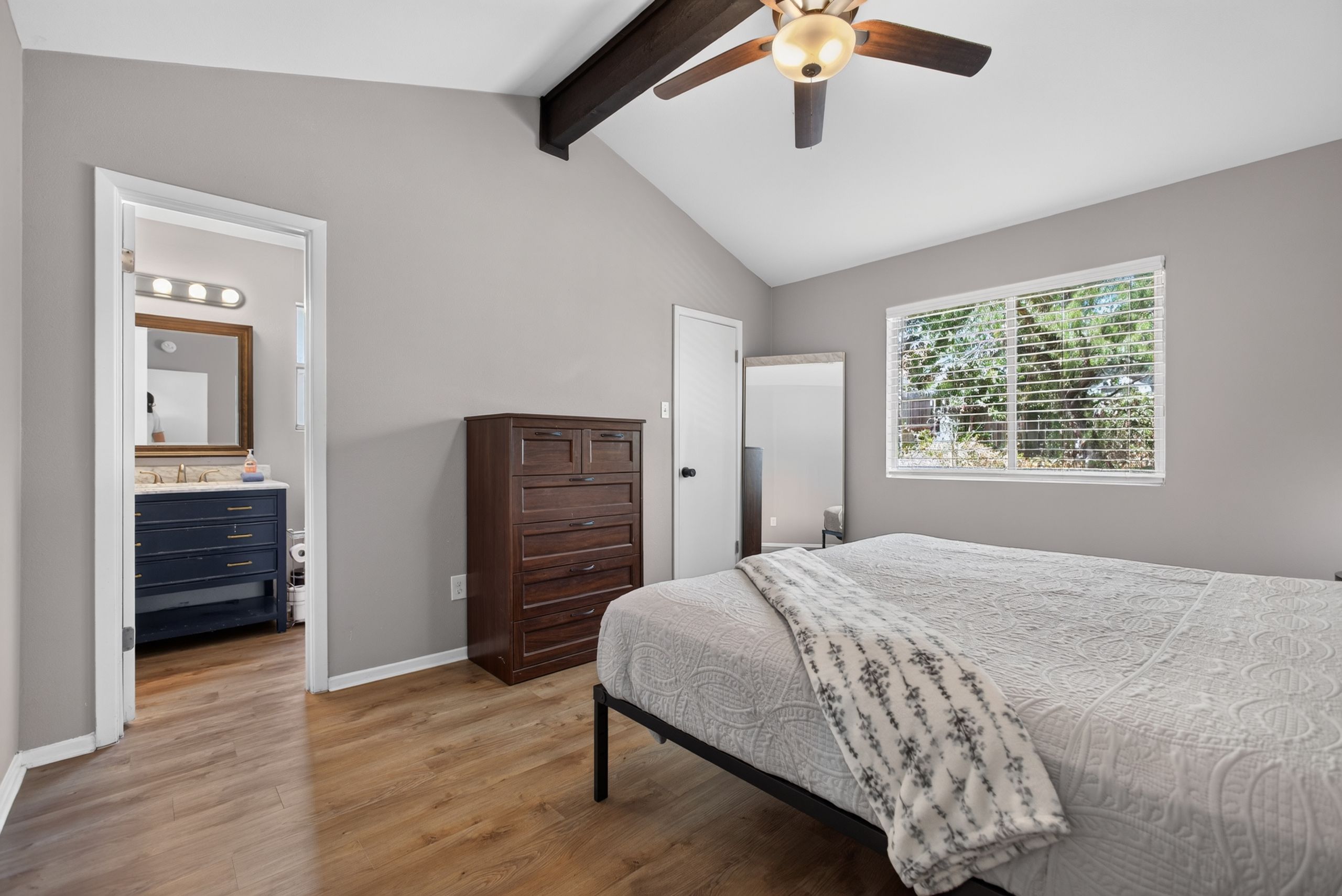 A bedroom with a bed, a dresser, a vanity with lights, a ceiling fan, and a window with blinds.