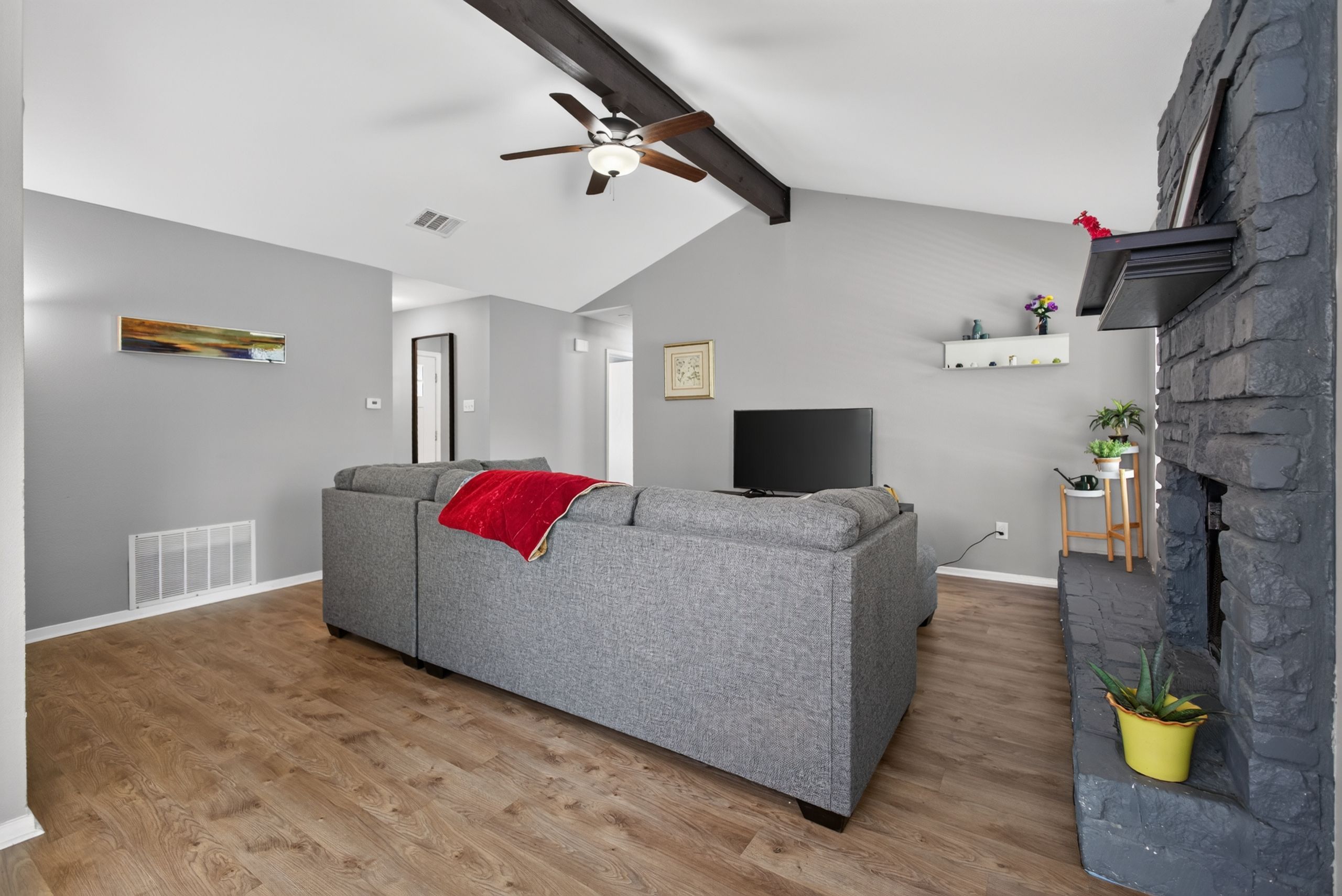 Un salon moderne avec un canapé gris, un plaid rouge, un ventilateur de plafond, un téléviseur à écran plat et une cheminée en pierre.