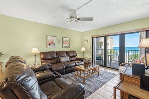 Ein Wohnzimmer mit einer braunen Ledersofa, einem Glastisch und einem Deckenventilator. An den Wänden hängen zwei gerahmte Bilder, und durch eine Schiebetür blickt man aufs Meer.