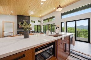 A modern kitchen with a large island, wooden cabinetry, and a view of a balcony with a scenic landscape. - The Laurel & Sky ~ Smoky Mountains (Bryson City)