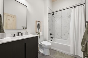 Une salle de bain moderne avec des toilettes blanches, une baignoire avec un rideau de douche et un meuble-lavabo doté d’un miroir et d’un porte-serviettes.