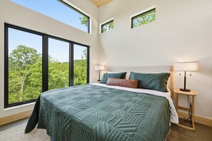 A bedroom with a large bed, two bedside lamps, and a view of trees through the windows. - The Laurel & Sky ~ Smoky Mountains (Bryson City)