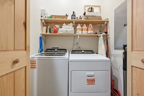 Une buanderie équipée d’un lave-linge et d’un sèche-linge, d’une étagère contenant des produits de nettoyage et d’un aspirateur rouge.