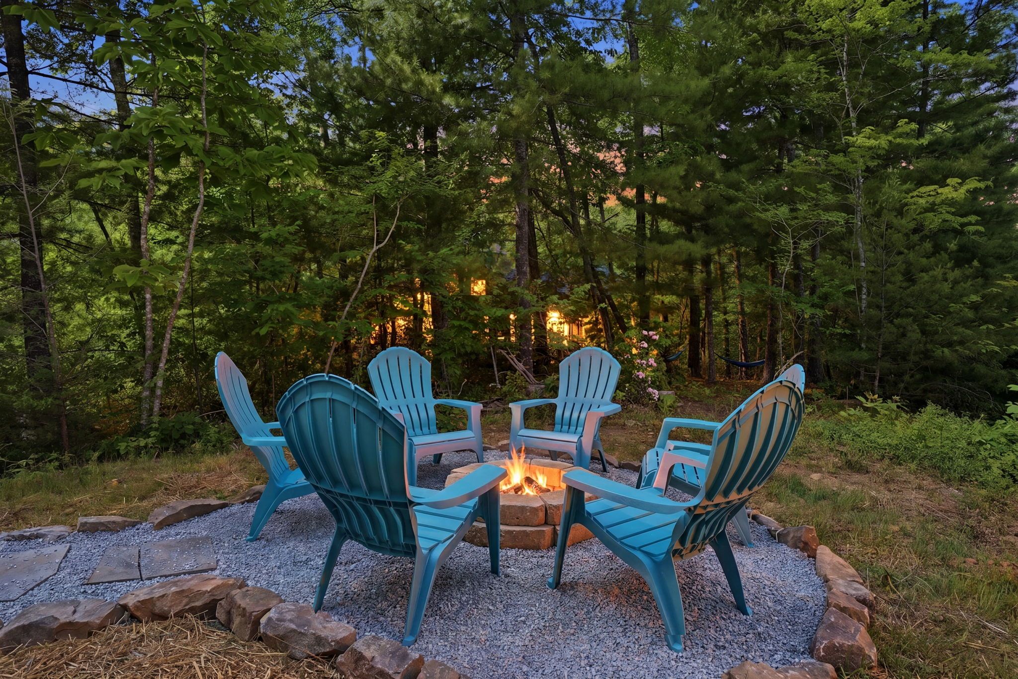 A fire pit with four Adirondack chairs arranged around it, set on a gravel surface with trees in the background.