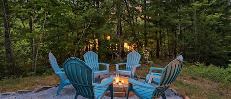 A fire pit with four Adirondack chairs arranged around it, set on a gravel surface with trees in the background.