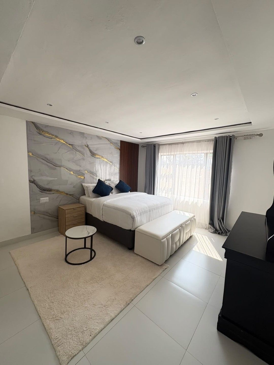 A modern bedroom with a bed, bedside tables, a desk, and a large window with curtains.