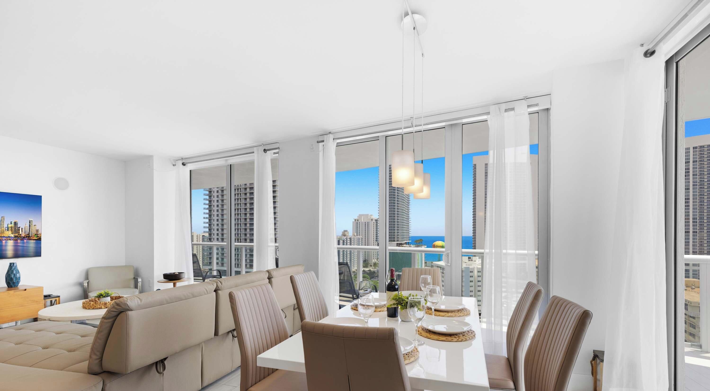 A modern living room with a sofa, dining table, and chairs. There is a large window with a city view.