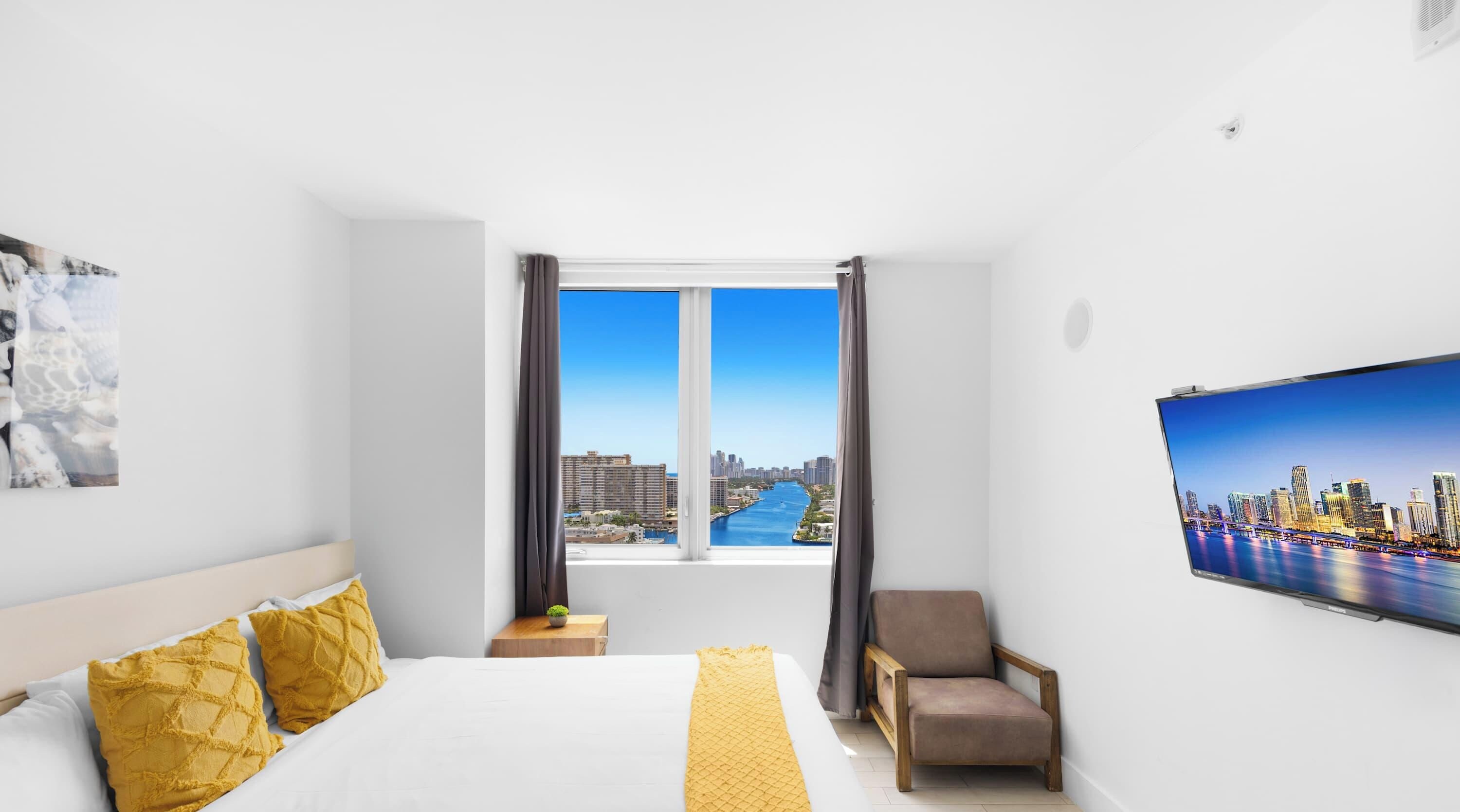 A hotel room with a large bed, a chair, a TV, and a window with a city view.