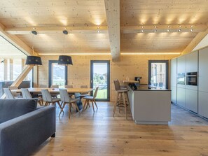 Ein modernes Interieur mit Holzbalkendecke, einem Essbereich mit Stühlen und Tisch, einer Küche mit Geräten und Blick ins Freie.