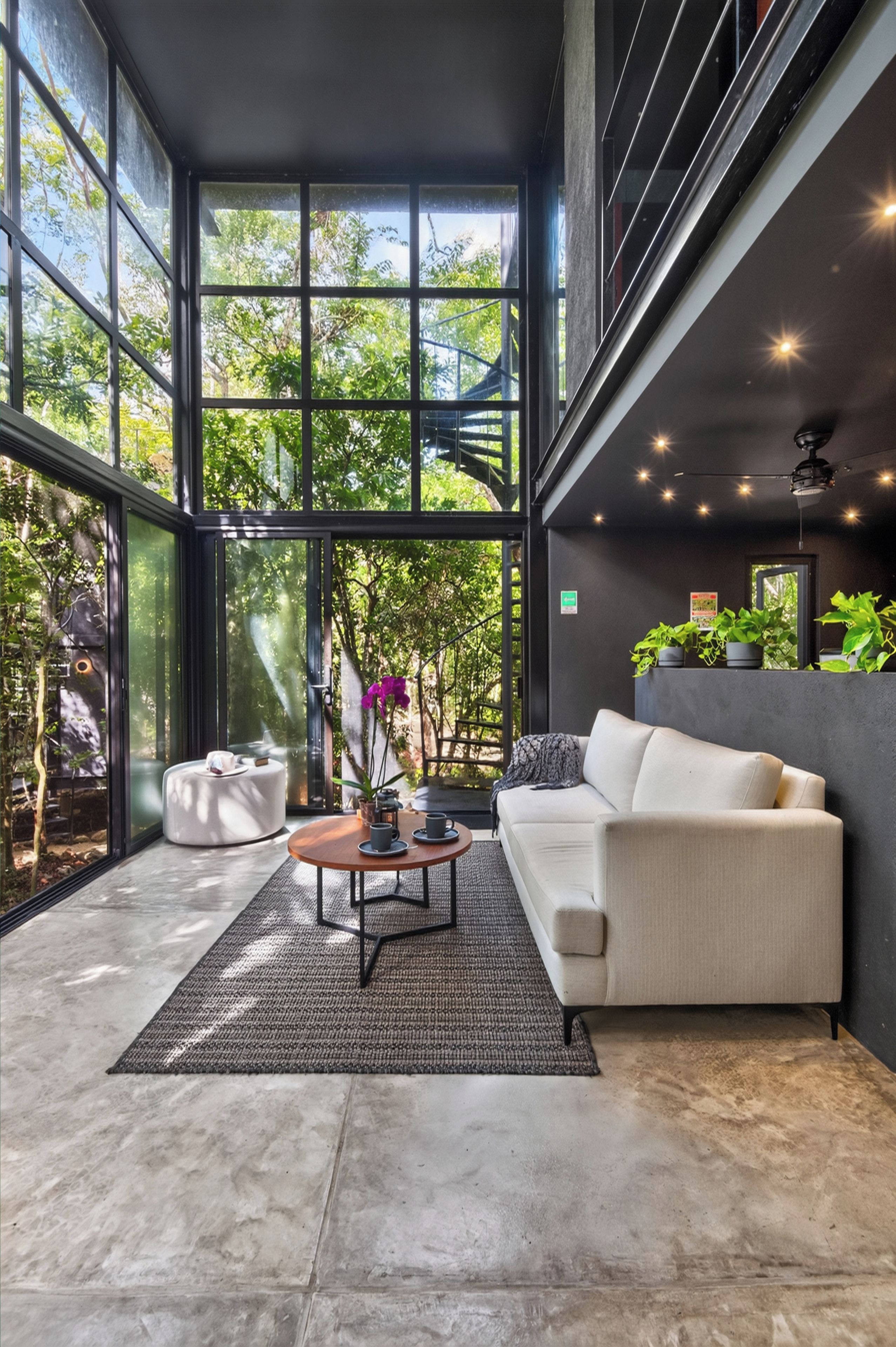 Un intérieur moderne avec une grande baie vitrée, un canapé blanc, une table basse et une petite table sur laquelle se trouvent des tasses et un vase avec une fleur.