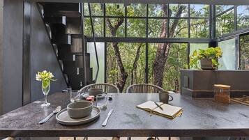 Un comedor moderno con una mesa lista para dos personas, un jarrón con flores y vistas a árboles a través de amplias ventanas.