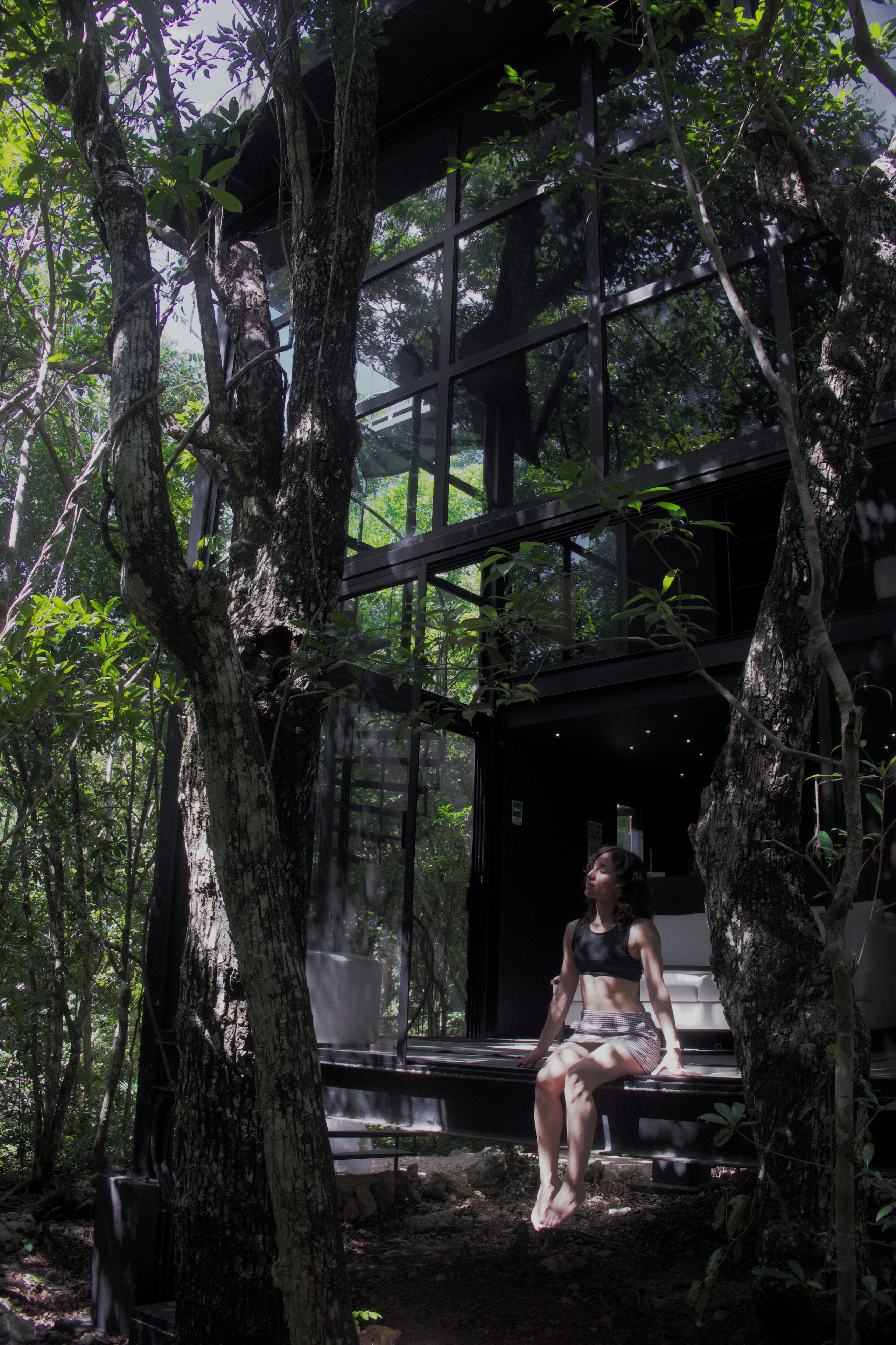 A woman sitting on a bench in a forested area with a glass structure in the background.