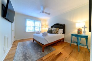 A bedroom with a bed, a ceiling fan, a window with blinds, a bedside lamp, and a small table with a lamp.