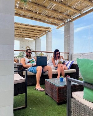 Un área de estar al aire libre con mobiliario de mimbre y una mesa, bajo una estructura de techo de paja.