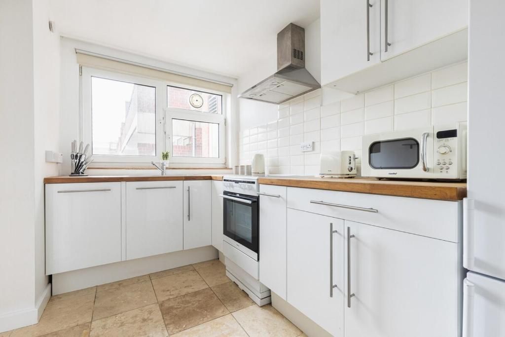 A modern kitchen with white cabinets, a built-in oven, a microwave, and a window with blinds.