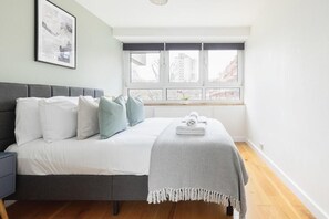 A bedroom with a bed, a bedside table, a window with a view of buildings, and a framed picture on the wall. - Central London Cosy 1BR Flat | Near Battersea Park (London)