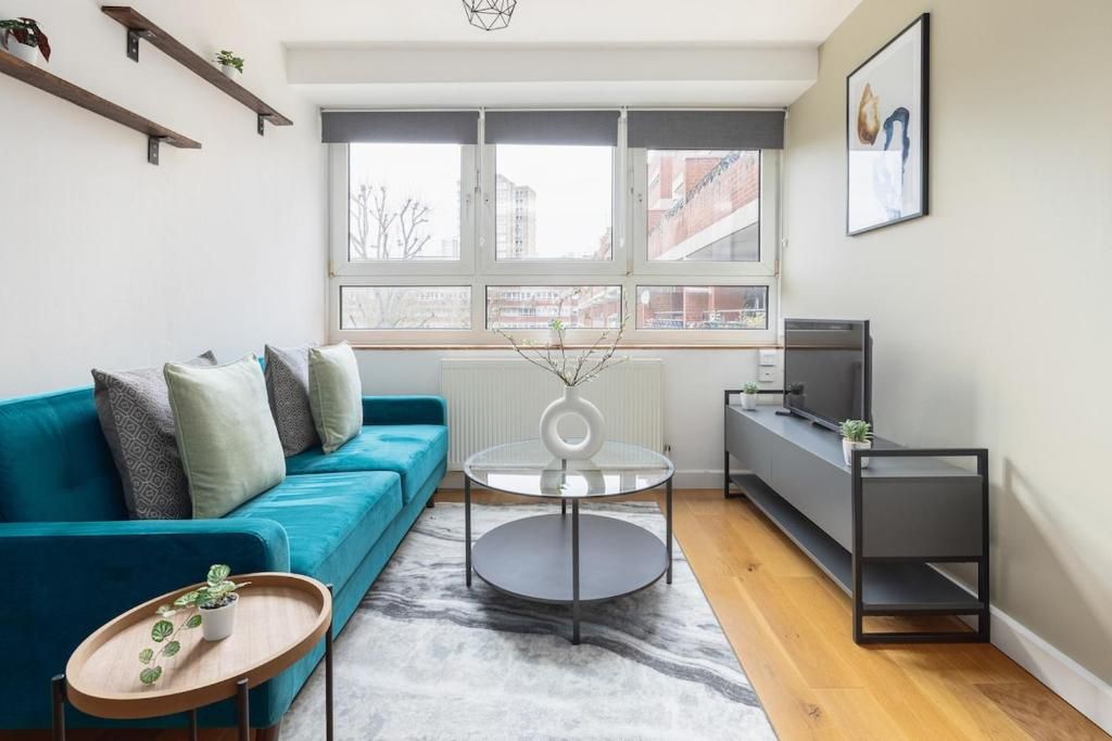 A modern living room with a teal sofa, a glass coffee table, a grey rug, and a view of the city through large windows.