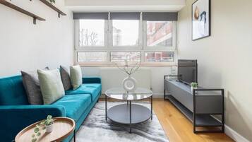 A modern living room with a teal sofa, a glass coffee table, a grey rug, and a view of the city through large windows.