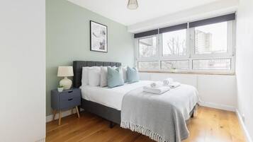 A bedroom with a bed, bedside table, lamp, and a window with a view of buildings.