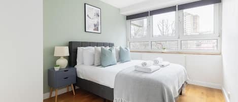 A bedroom with a bed, bedside table, lamp, and a window with a view of buildings.