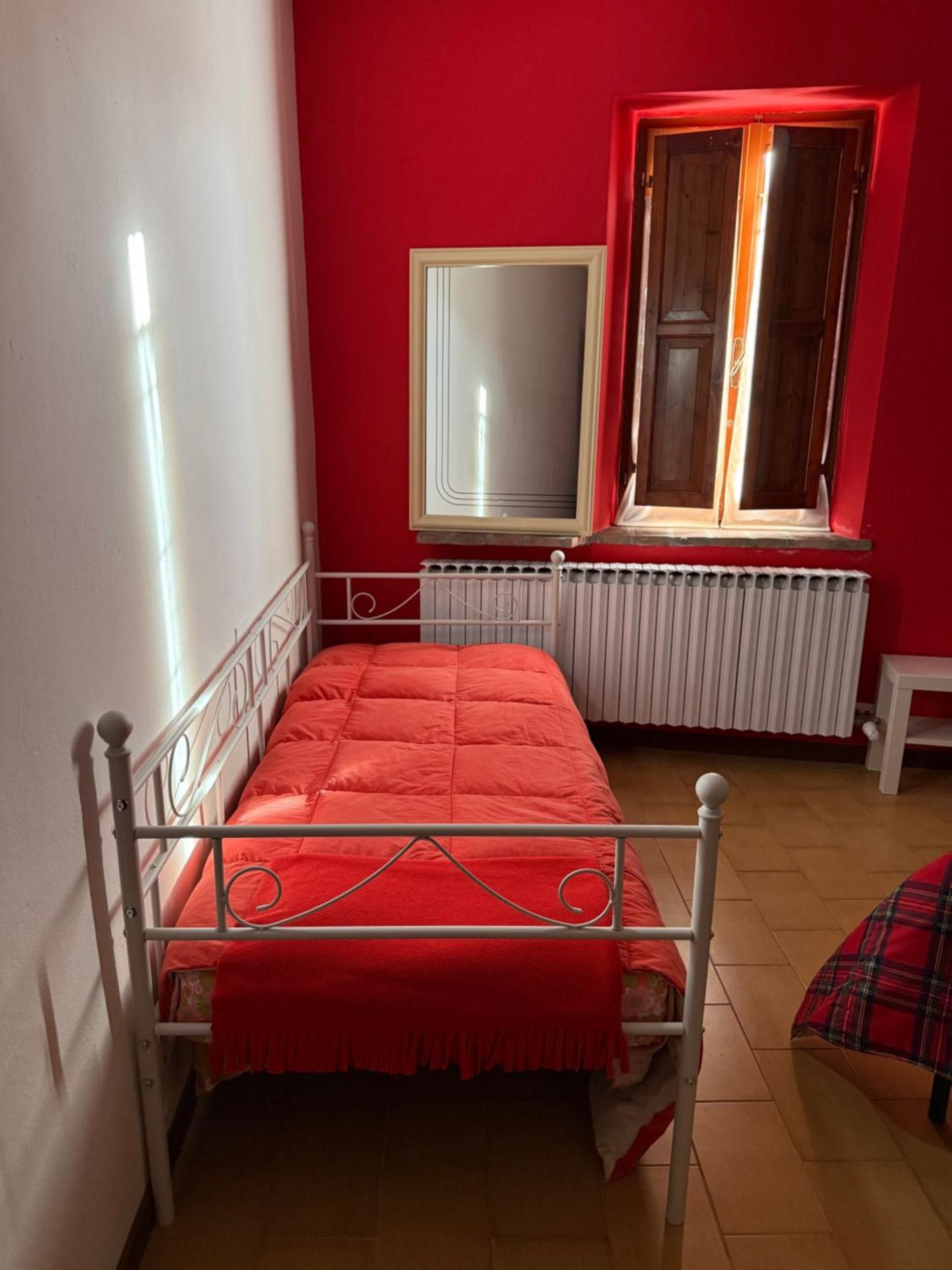 A single-bed room with a red wall, a window with curtains, a radiator, and a mirror.