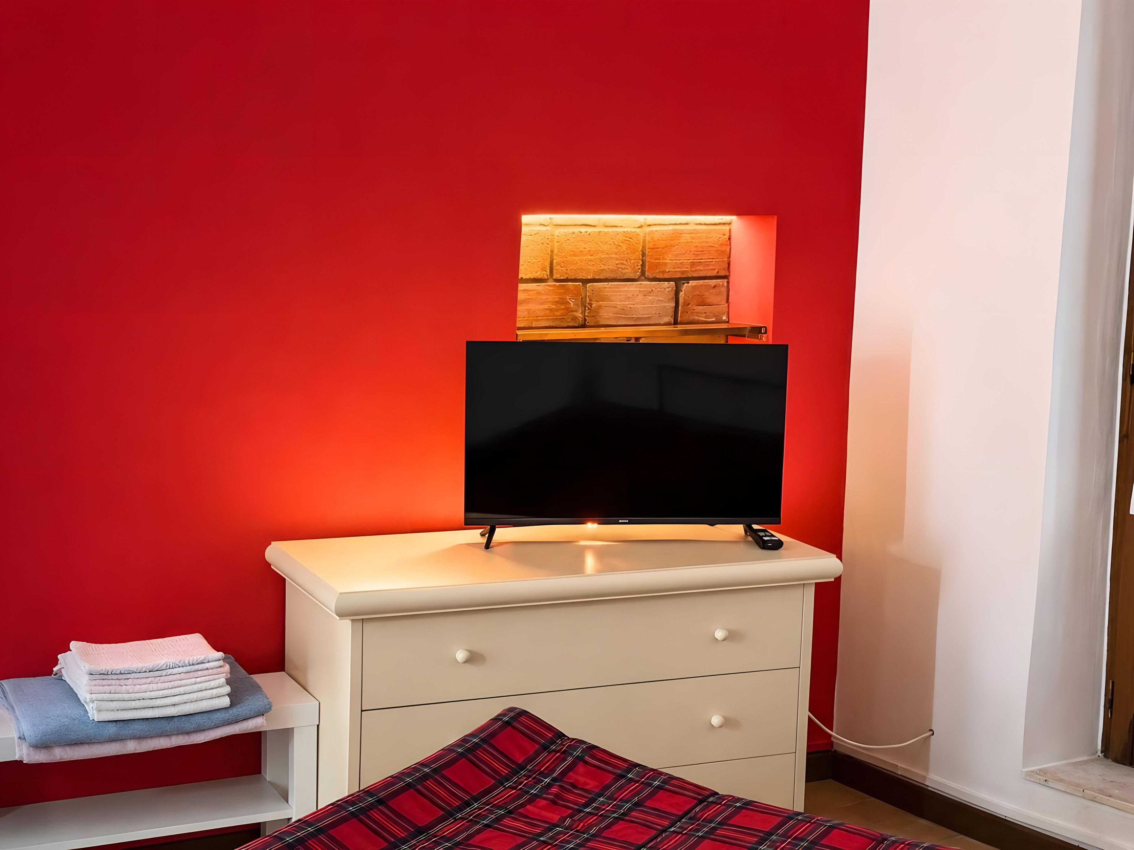 Ein Zimmer mit Fernseher auf einer weißen Kommode, einer roten Wand und einem Fenster.