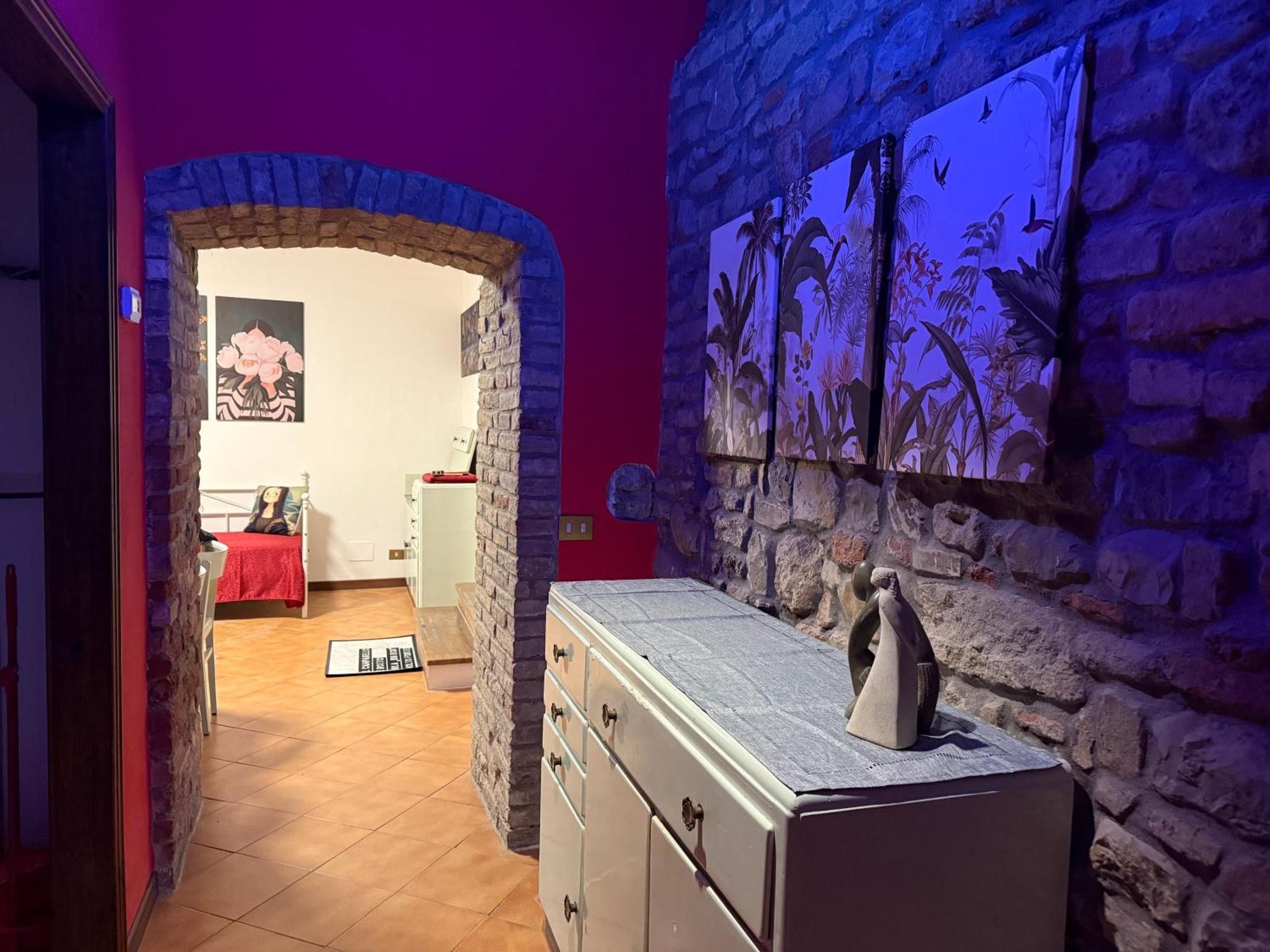 A room with a stone wall, a bed with a red cover, a dresser, and a wall-mounted shelf with a sculpture.