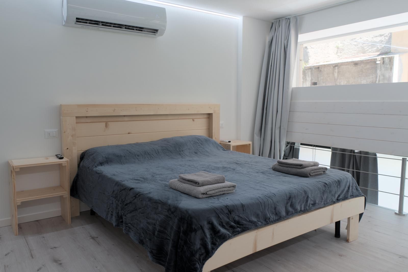 A bedroom with a wooden bed, a nightstand, a bedside table, a window with curtains, and an air conditioning unit.