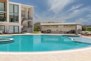 A modern pool area with a clear blue pool, a covered seating area with outdoor furniture, and a multi-story building with balconies.